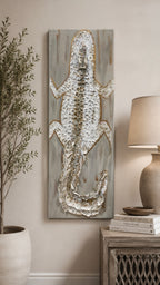 A vertical canvas wall art piece featuring a textured, raised alligator design in metallic silver and gold tones over a distressed gray wood-grain background, styled in a home decor setting next to a plant and a lamp.